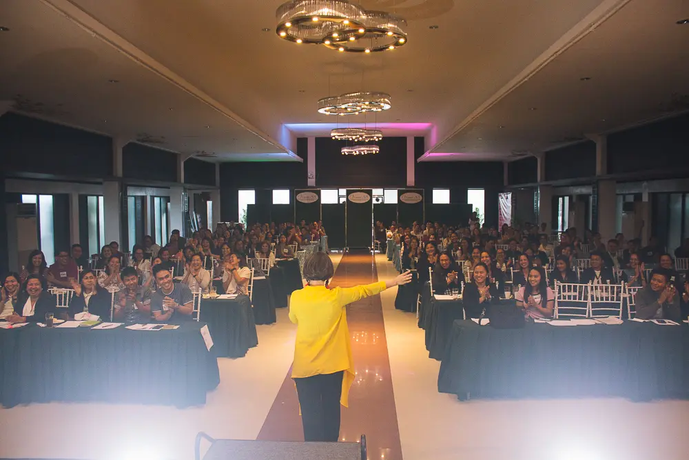 A woman in a yellow blouse standing on stage and receiving a round of applause from the crowd.