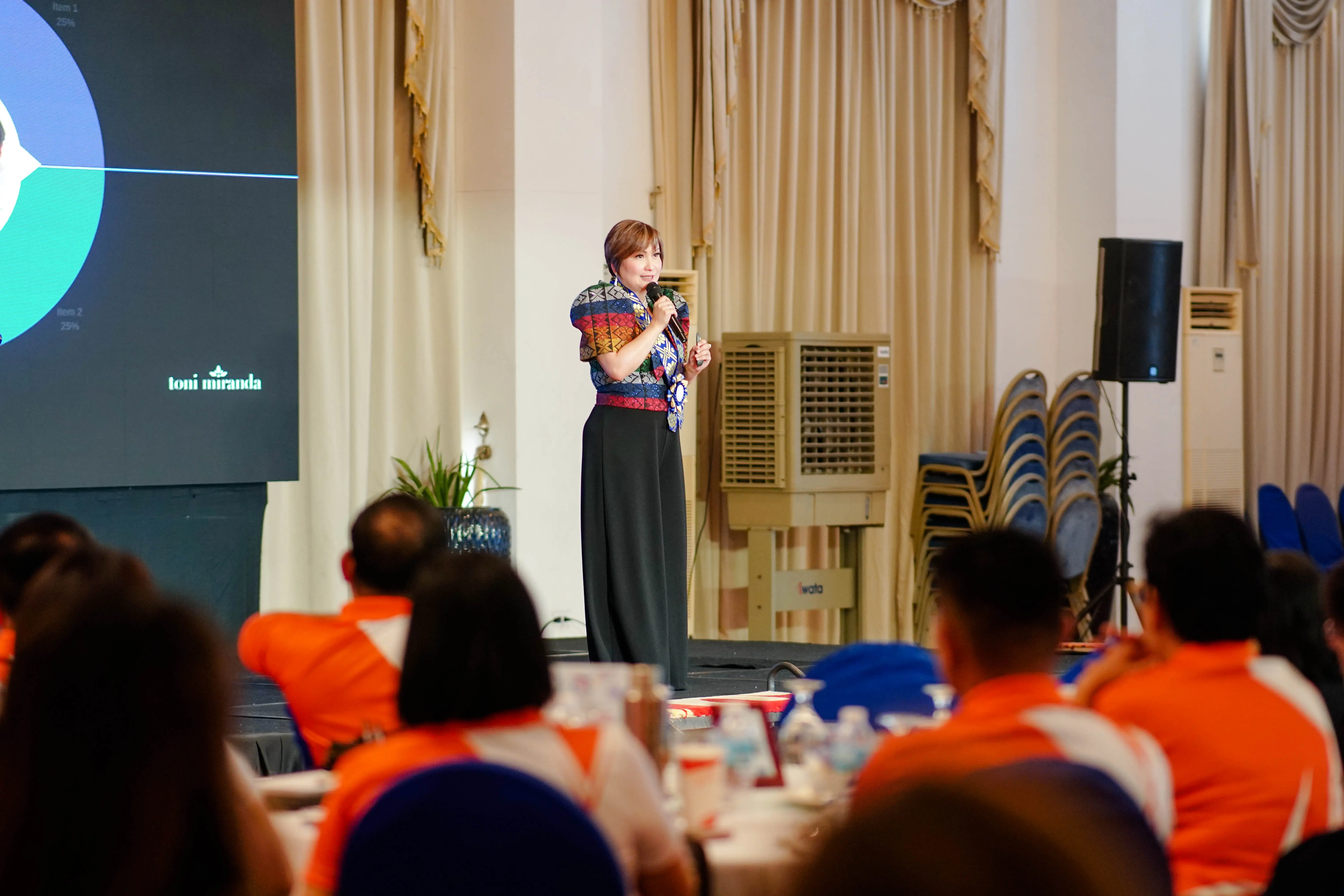 A woman in a modern Filipiniana, sharing a speech in front of an audience.