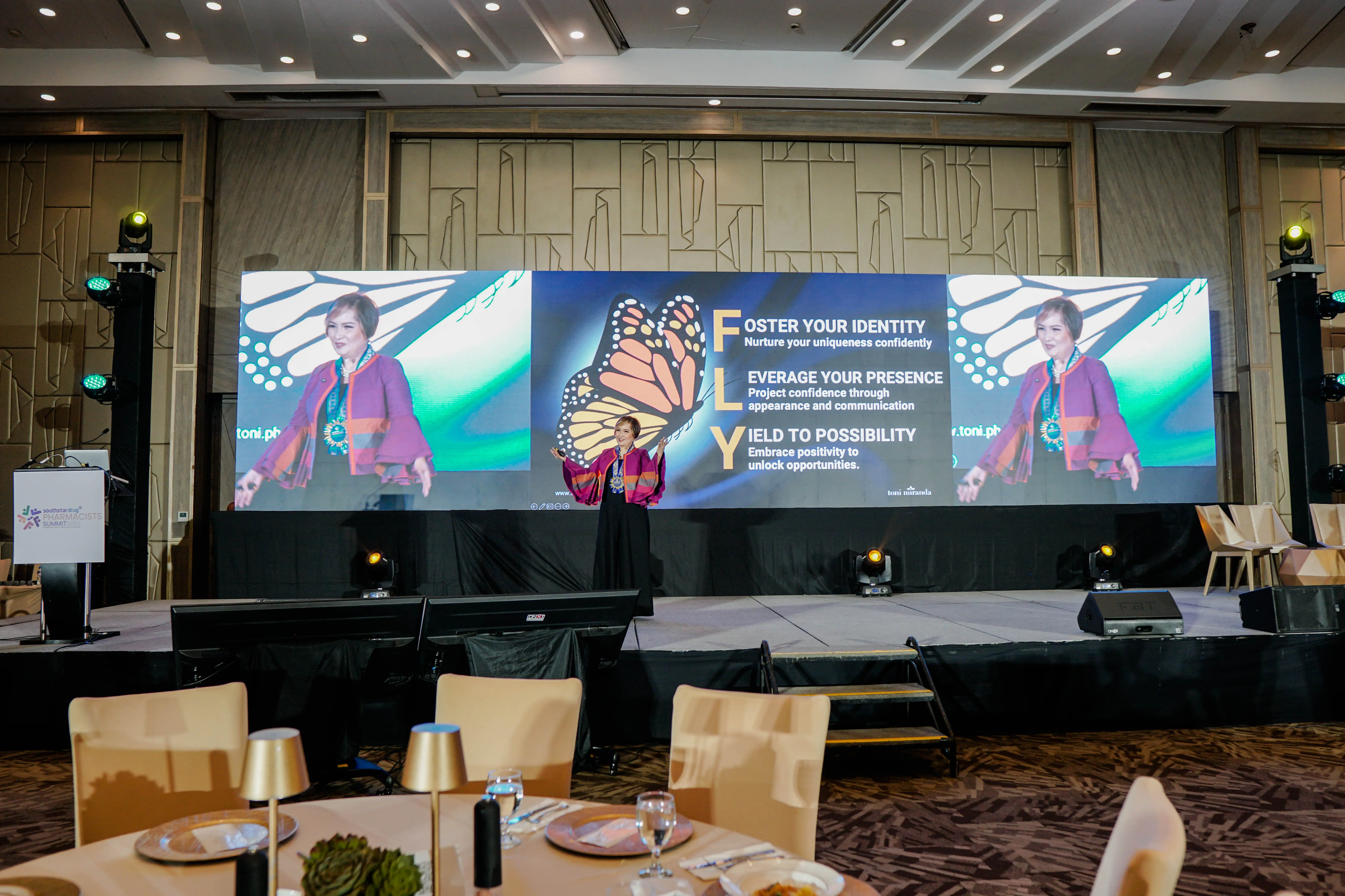 A woman in purple clothing talking on stage in a modern event setting.