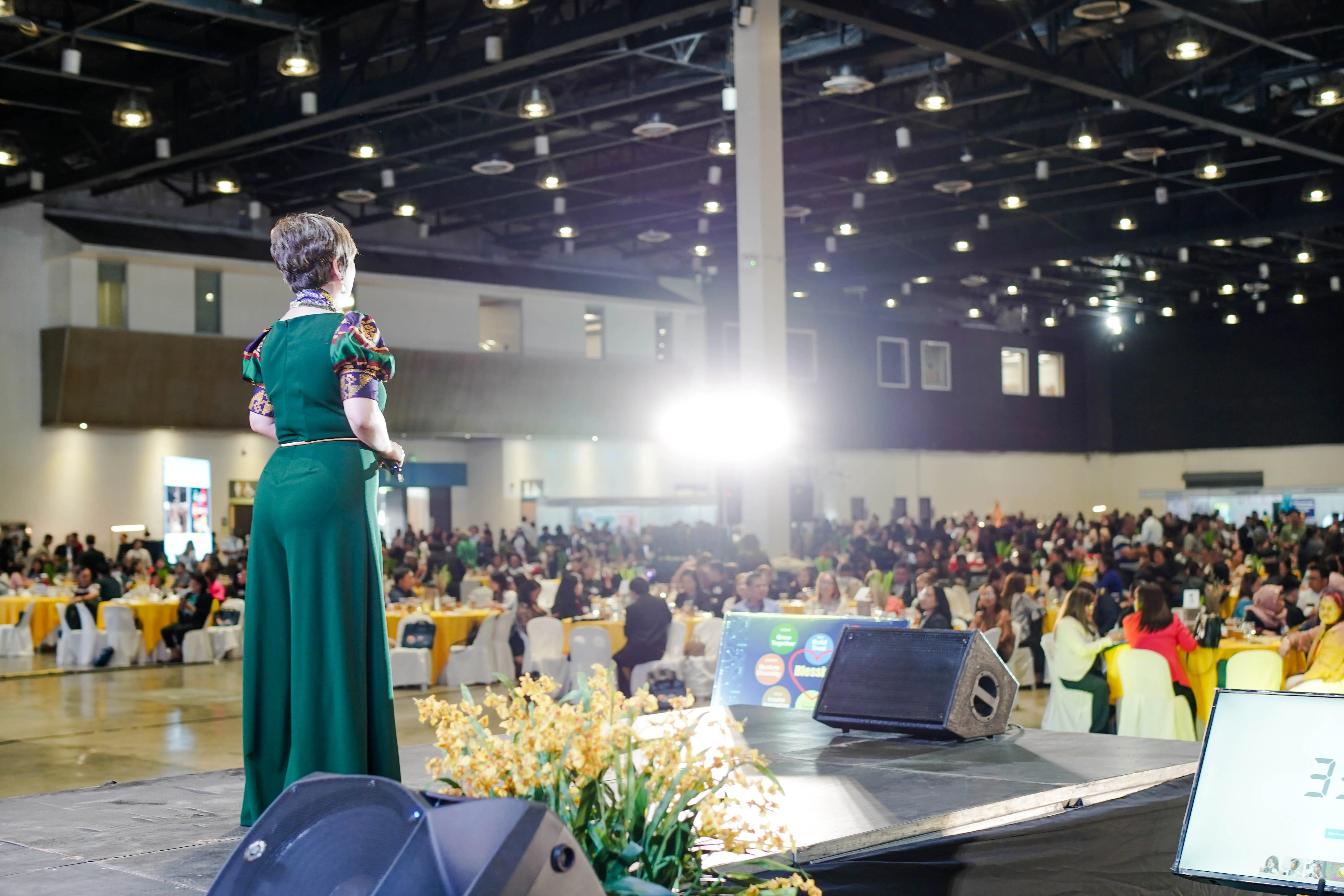 A woman in a green dress talking on stage before a big crowd.