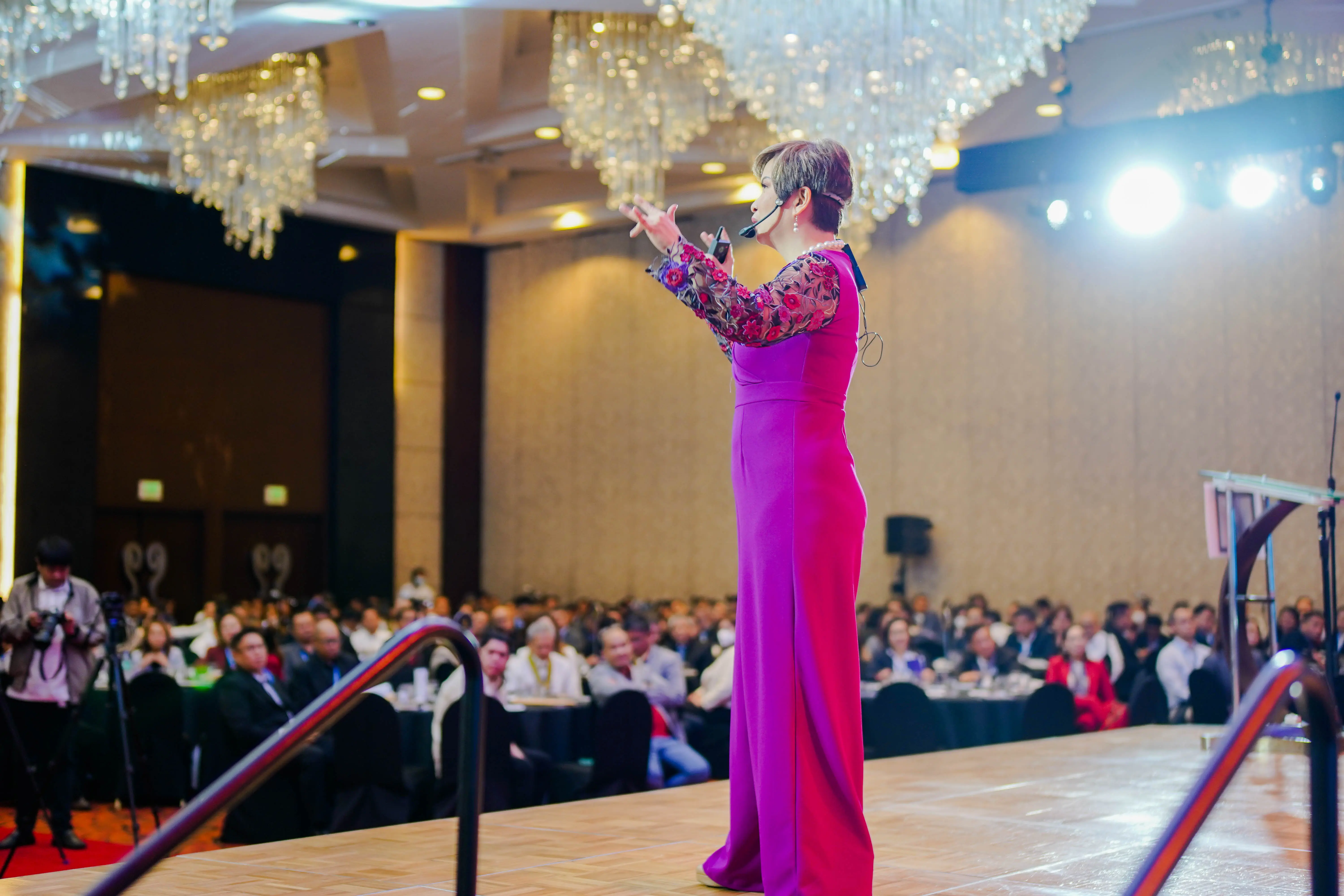 A woman in a bright pink dress speaking on stage in front of a big crowd.