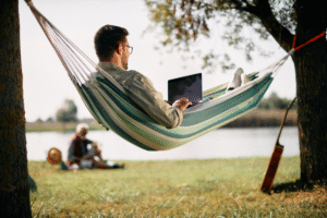 A young man lounging on a hammock while using a laptop.