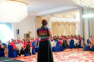 Toni Miranda wearing a multicolored bolero top and long skirt, speaking in front of a seated audience in an event