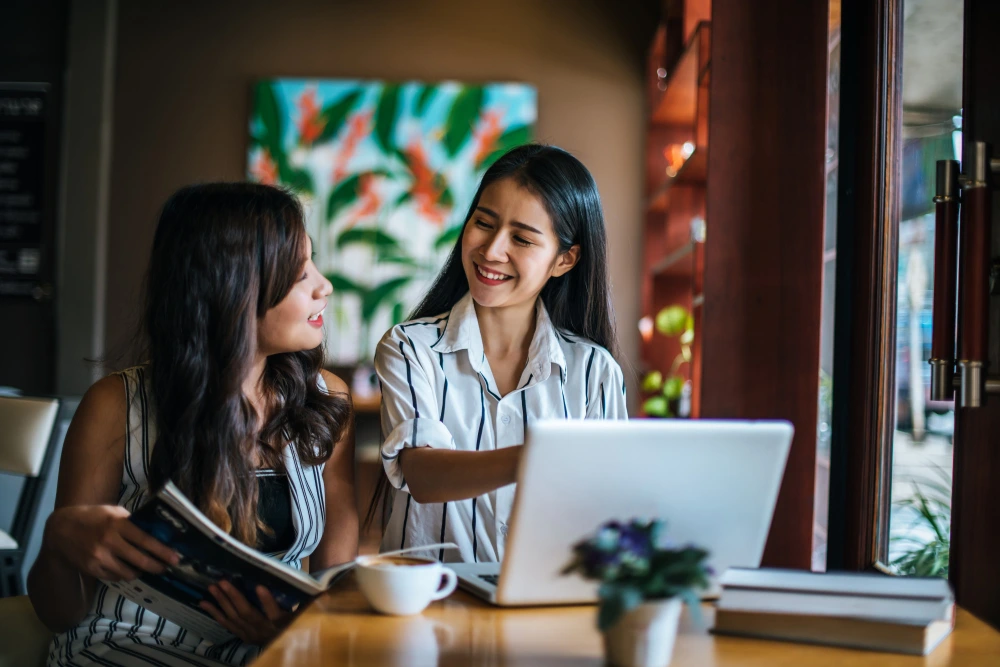 Two women in a cafe share a friendly moment, one holding a book, the other working on a laptop.