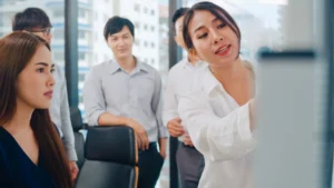 A woman in a white shirt writes on a whiteboard during a team meeting while colleagues watch attentively.